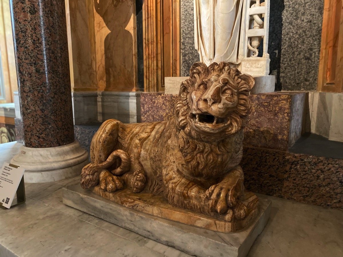 An alabaster carved lion reclining, its front paws crossed, mouth open