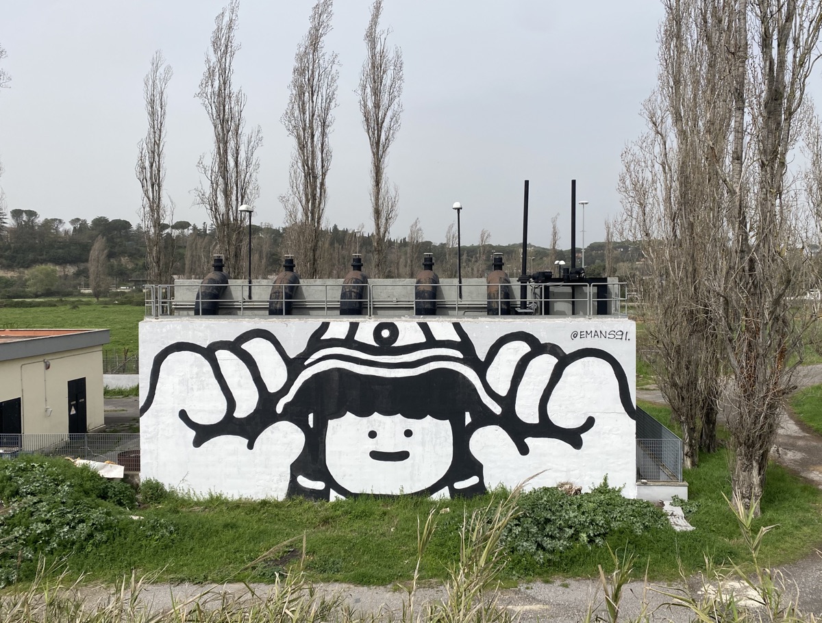 A black and white mural of a face beneath a bucket hat painted on a large plumbing structure at a sewage farm