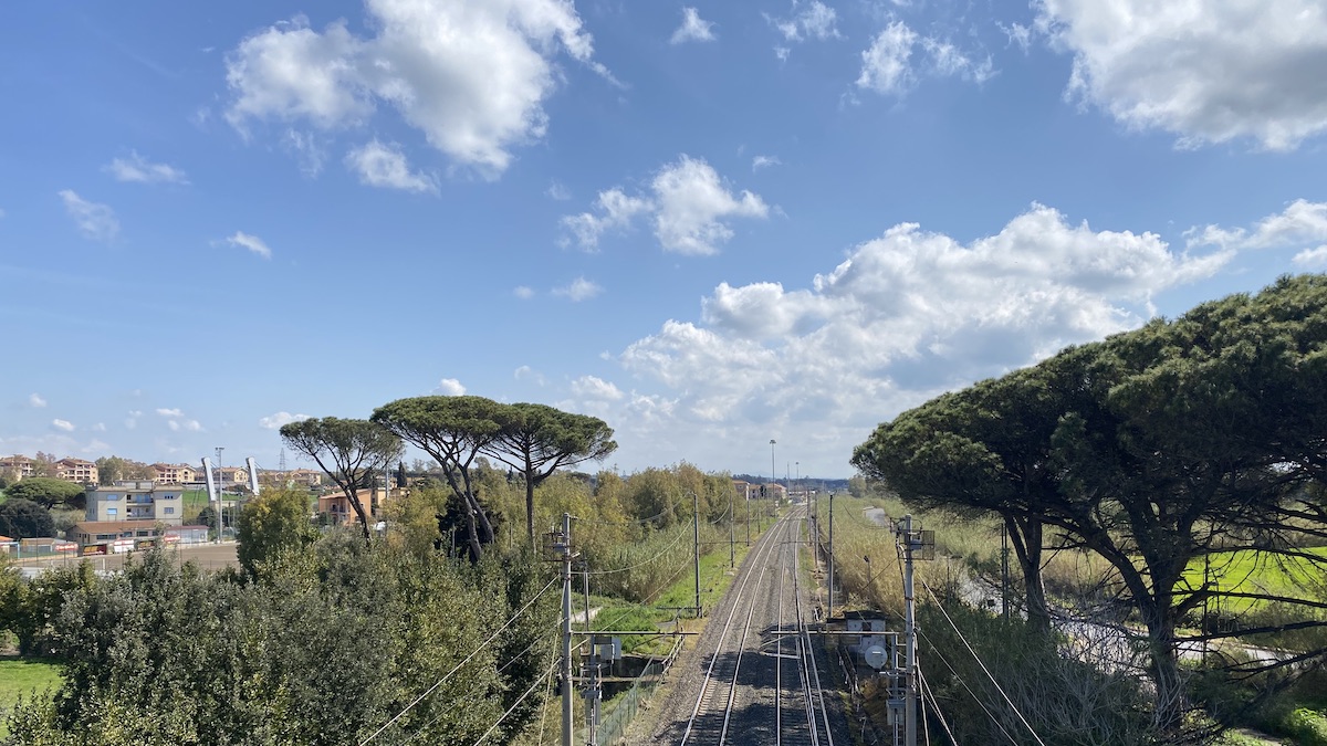 Railway tracks recede into the distance, bordered by umbrella pines and with fluffy clouds overhead in a blue sky