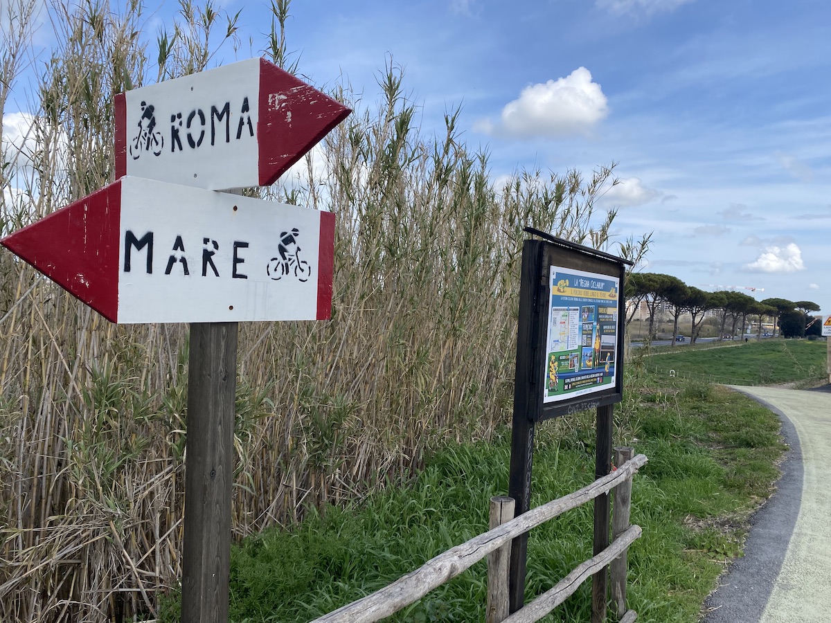 A signpost on the cycle path outside the twon of Fiumicino. Pointing left, a stencil of a cyclist with a backpack, to the sea, pointing right, to Rome.