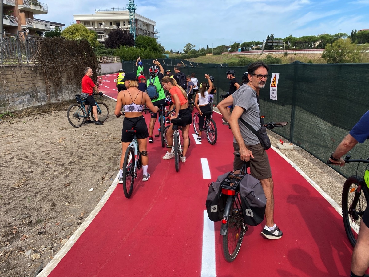 A group of cyclists seen from behind on a newly surfaced red cycle path with some apartment buildings on the left and a large road on the left.