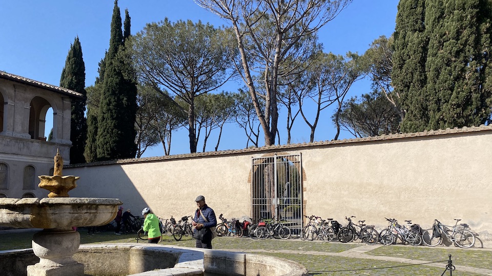 Lots of bicycles parked against the wall of the old monastery with a fountain in the foreground