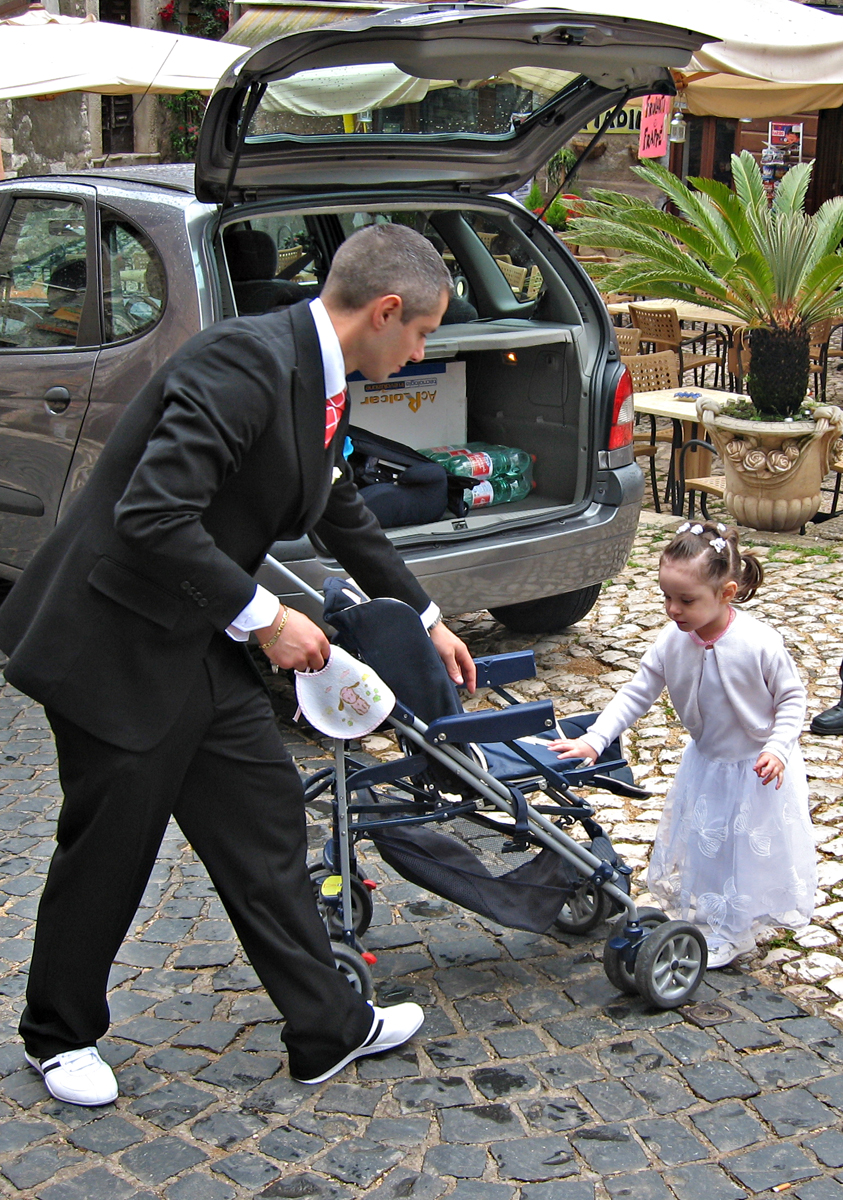A young man wearing a sharp suit with white trainers coaxes a young girl into a baby carriage. The girl may be dressed for first communion, though she seems a bit young