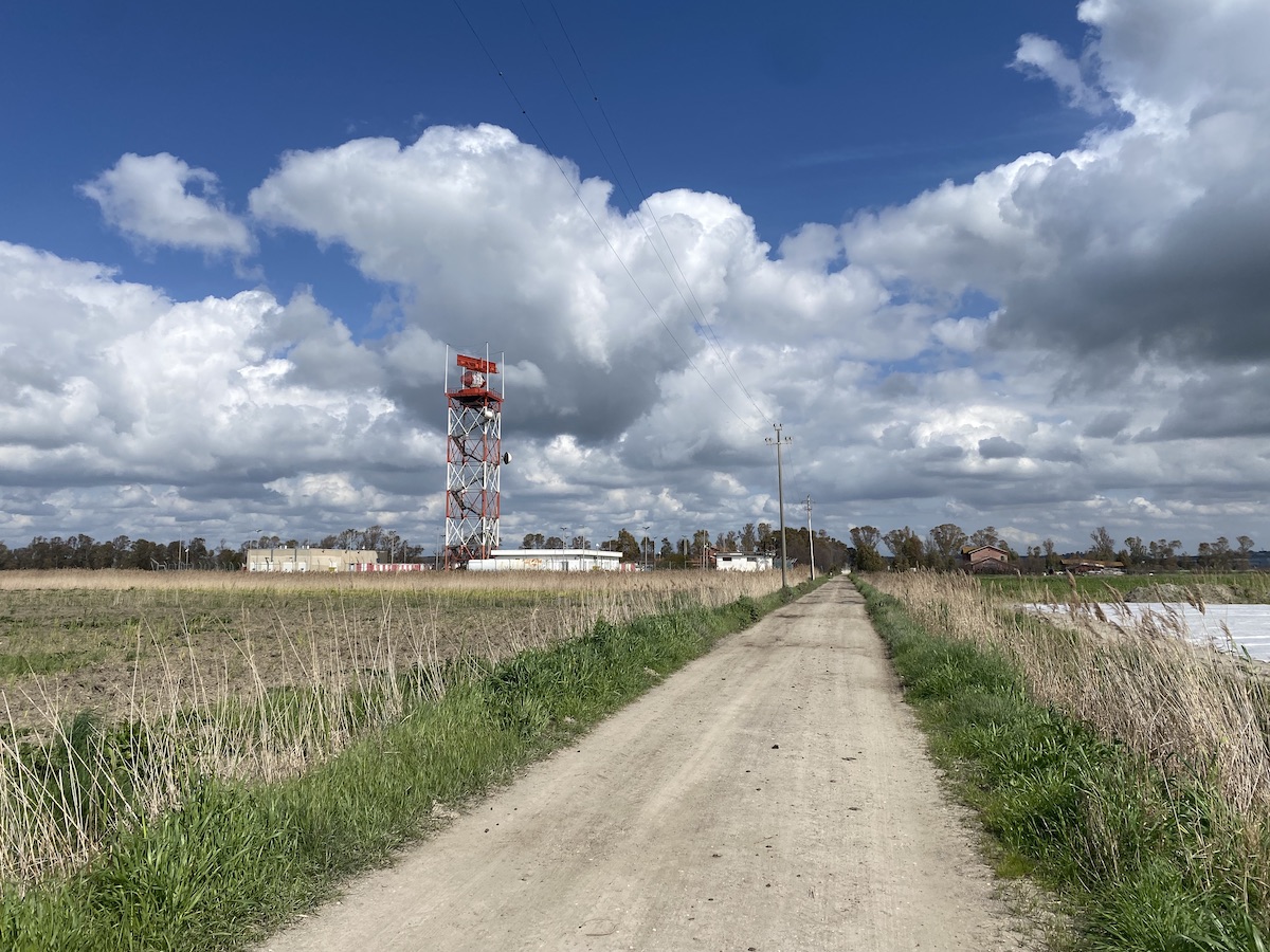 A gravel path between vegetable fields recedes into the distance. On the left6 is the red and white radar tower for the airport at Fiumicino. The blue sky is full of silvery clouds.