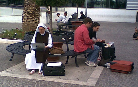 A nun, sitting on a park bench with her rolling suitcase on the ground beside her, working on a laptop that is on her lap surfing.jpg