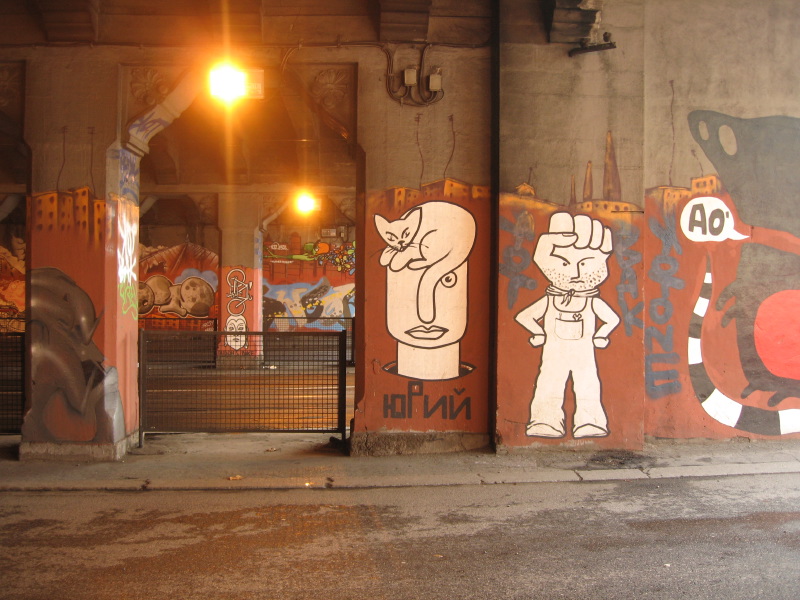 Graffiti under a bridge. A cat rests on a man's head and a man has a fist for a head